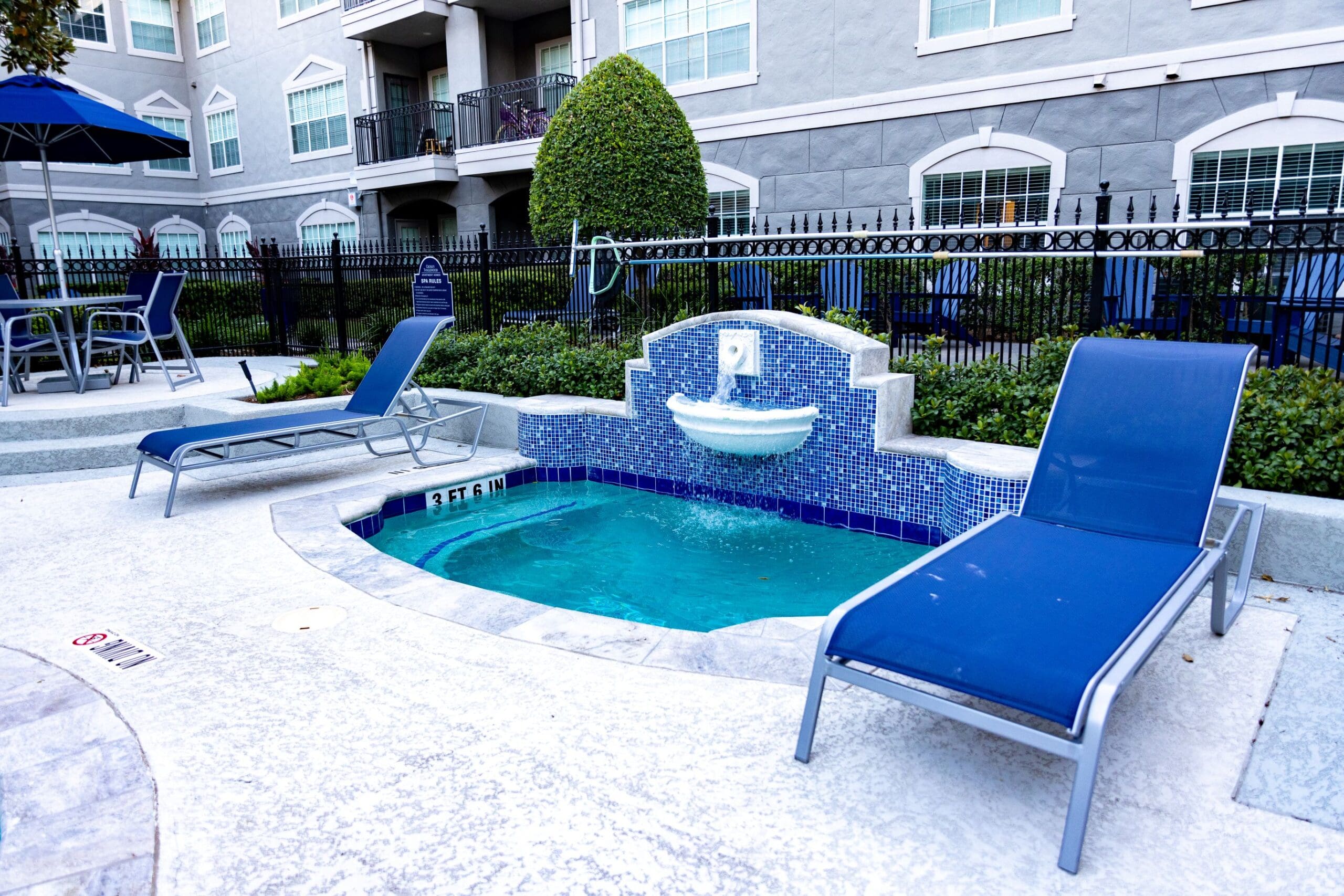 **Alt text:** Small spa or hot tub area at Crosley Tanglewood Apartment Homes, featuring a water fountain built into a blue tile mosaic wall, surrounded by lounge chairs and landscaping, with a gated apartment building in the background.