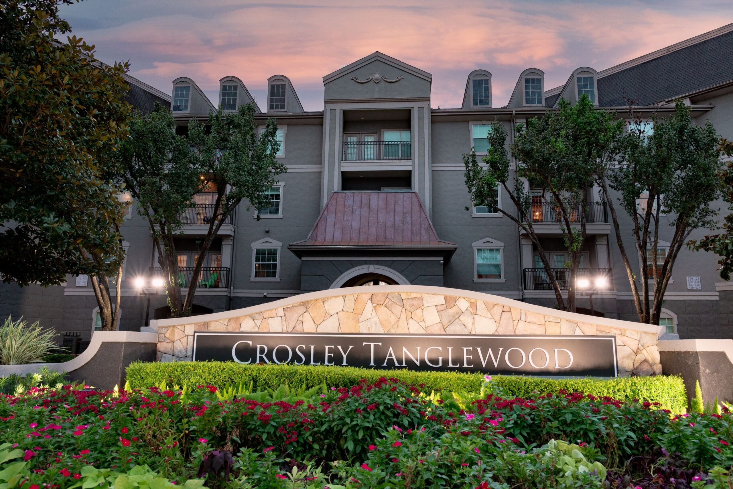 Elegant exterior view of Crosley Tanglewood Apartment Homes at dusk, featuring a stone monument sign surrounded by lush, colorful landscaping in the foreground, with a multi-story building and soft pink and blue evening sky in the background.