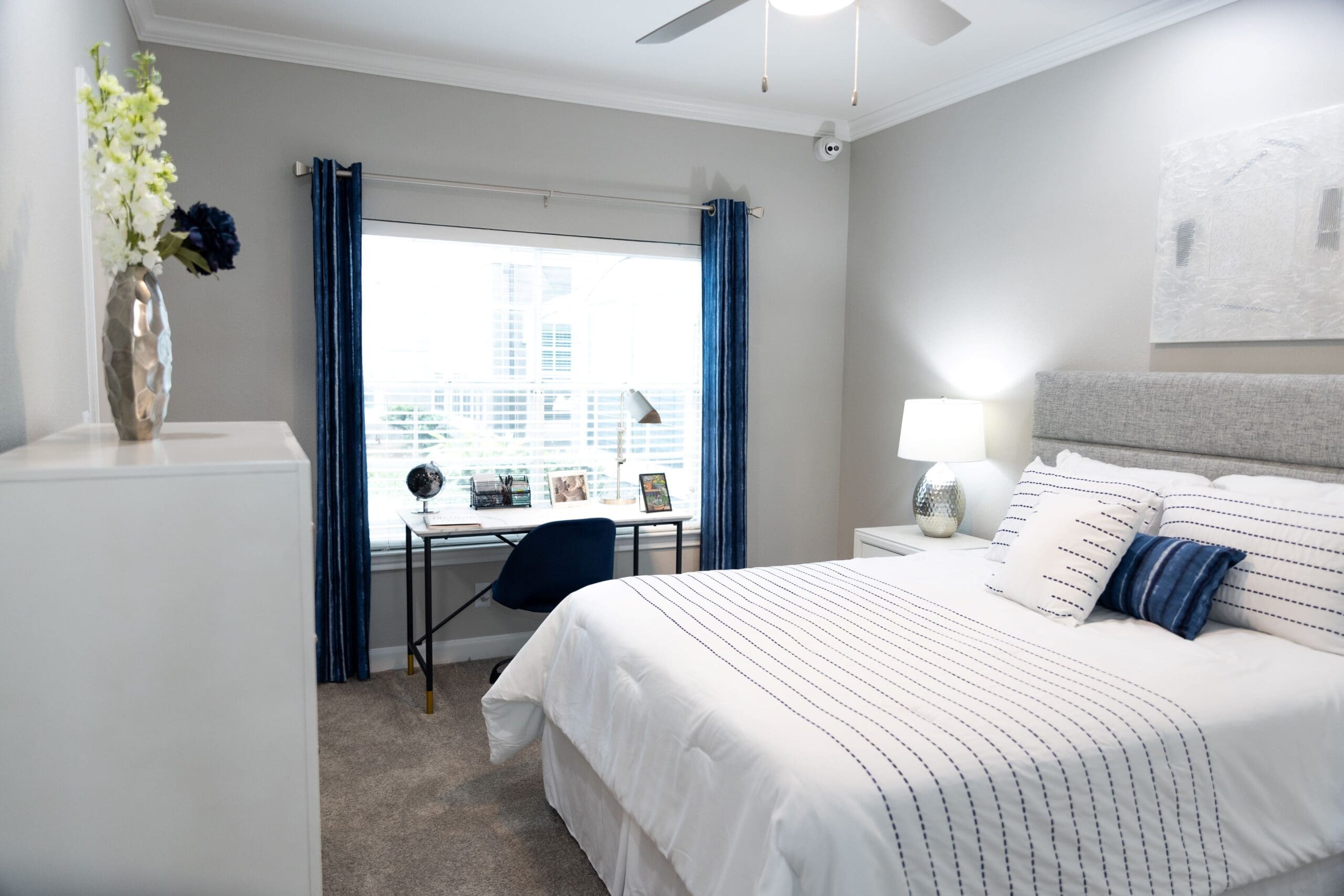 **Alt text:** A bright, neatly arranged bedroom featuring a full-sized bed with white bedding and blue accent pillows. A small white nightstand with a silver lamp is beside the bed. Opposite the bed is a minimalist desk placed by a large window with blue curtains, decorated with framed photos, a globe, and a desk lamp. A navy chair is tucked under the desk, and a tall white dresser with a decorative vase stands in the foreground.