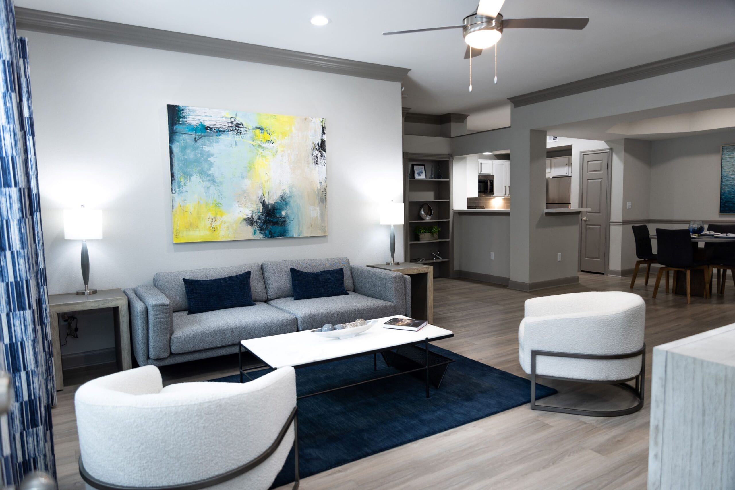 Apartments in Tanglewood, TX - Crosley Tanglewood - Spacious Living Area with Wood-Style Plank Flooring, Blue Area Rug, Grey Sofa, White Armchairs, White Coffee Table, Side Tables with Lamps, Wall Art, Ceiling Fan, and View of Dining Area and Kitchen.