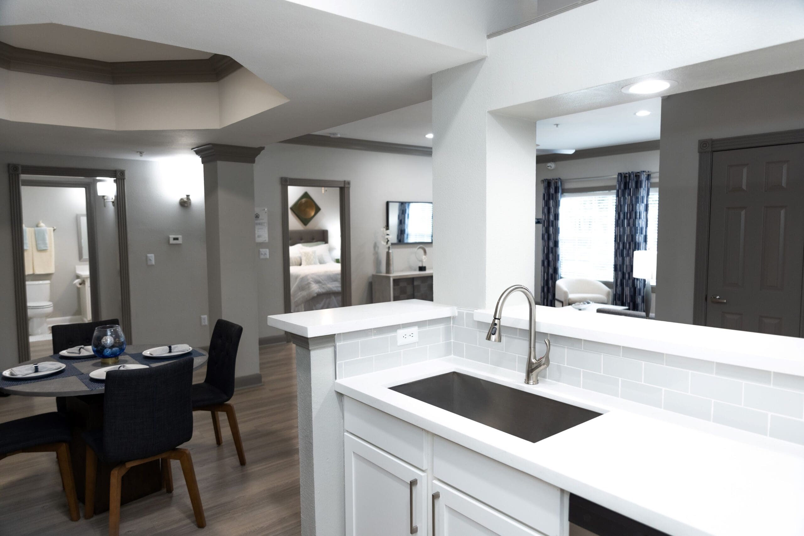 **Alt text:** View from a modern kitchen with a stainless steel sink and white cabinetry, overlooking a stylish apartment interior featuring a dining area with a round table and four dark upholstered chairs, and open sightlines to two bedrooms and a living room with blue curtains.
