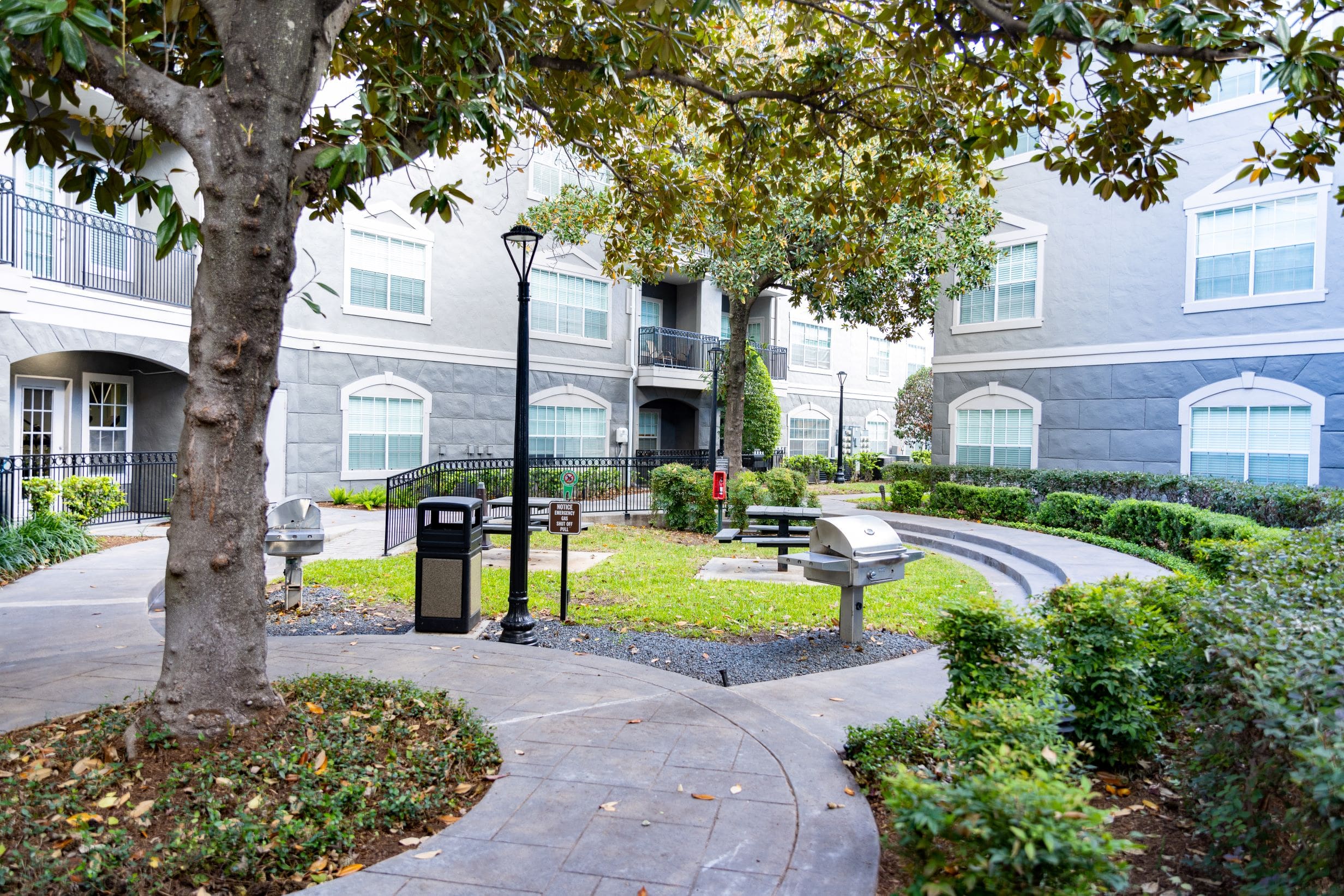 **Alt text:** A landscaped courtyard surrounded by gray apartment buildings featuring a communal outdoor space with two stainless steel barbecue grills, trash bins, picnic tables, benches, street-style lamps, and mature trees providing shade along the curved walkway.