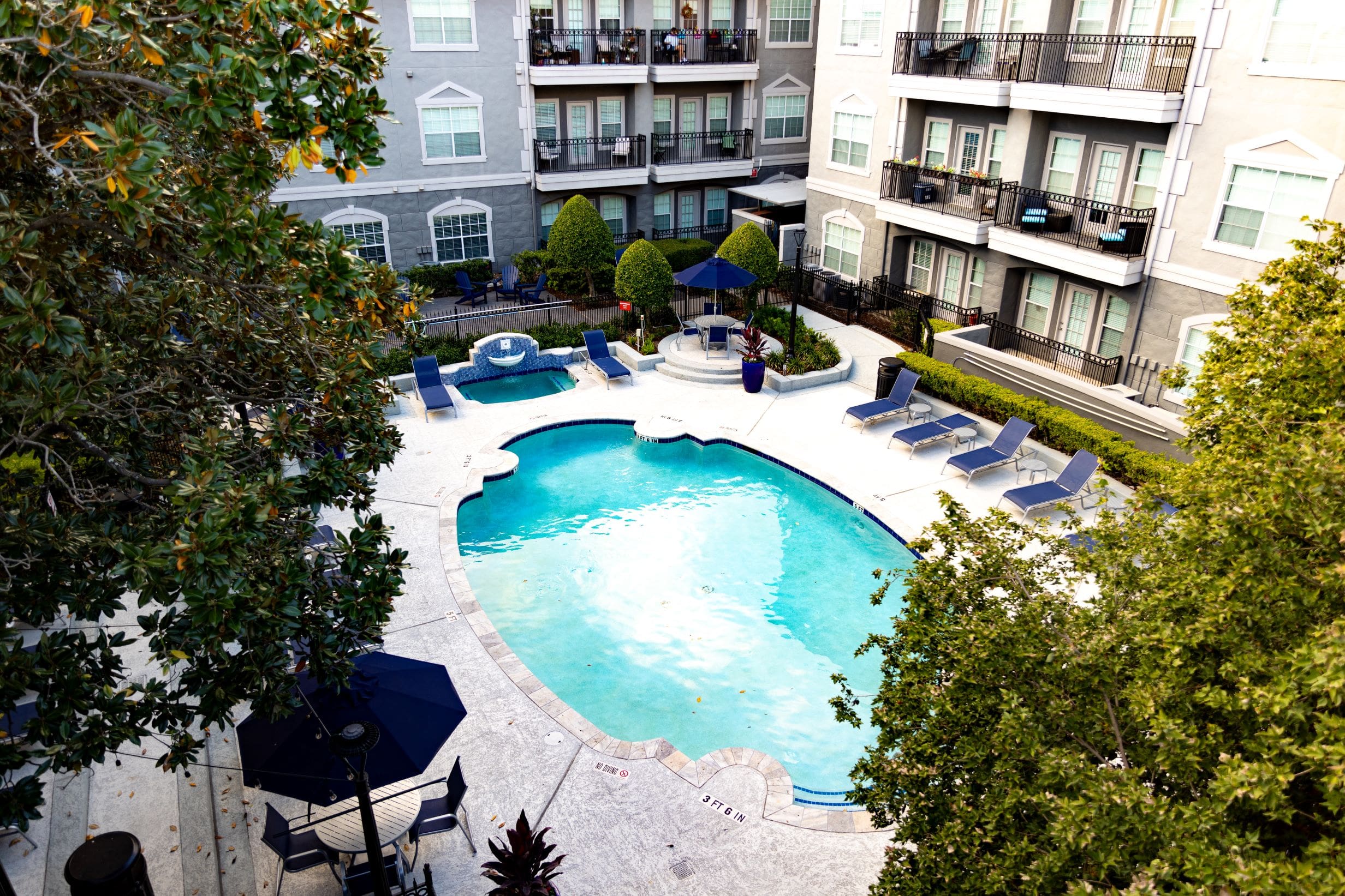 **Alt text:** Aerial view of a residential courtyard showcasing a swimming pool surrounded by lounge chairs, a hot tub with a decorative water feature, and umbrella-covered seating areas. The scene is framed by well-maintained landscaping and multi-story apartment buildings with balconies.