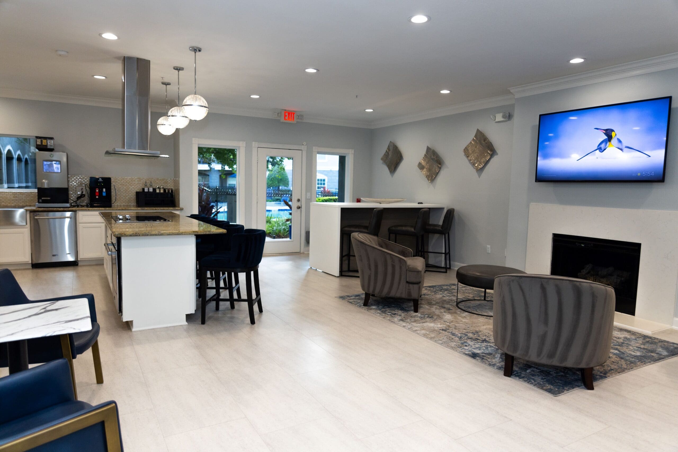**Alt text:** Modern clubhouse interior featuring a full kitchen with granite countertops, stainless steel appliances, and a central island with barstool seating. The space includes additional seating areas with cushioned chairs, a mounted TV above a fireplace, wall art, and glass doors leading to an outdoor area.