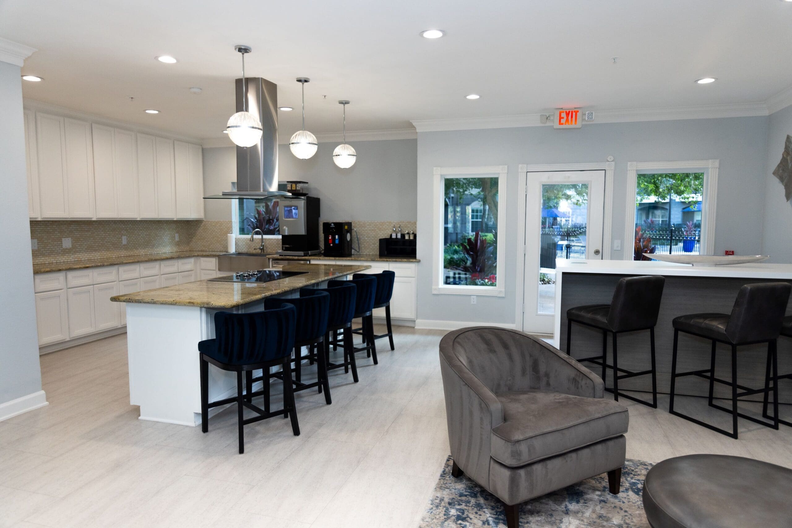 **Alt text:** Spacious communal kitchen and lounge area featuring a long granite island with stovetop and sink, surrounded by black barstools. The space includes modern pendant lighting, white cabinetry, stainless steel appliances, a coffee station, and gray lounge chairs near large windows and a door leading to an outdoor patio.