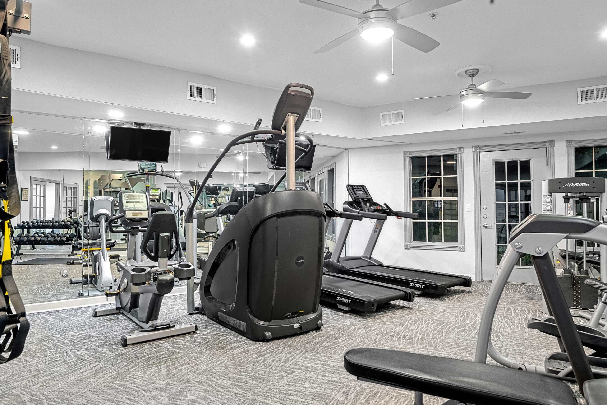 **Alt Text:** *Modern fitness center featuring a variety of cardio equipment including treadmills, a recumbent bike, and an elliptical machine. The space has full-length mirrors, ceiling fans, and a wall-mounted TV, providing a clean and well-lit workout environment.*